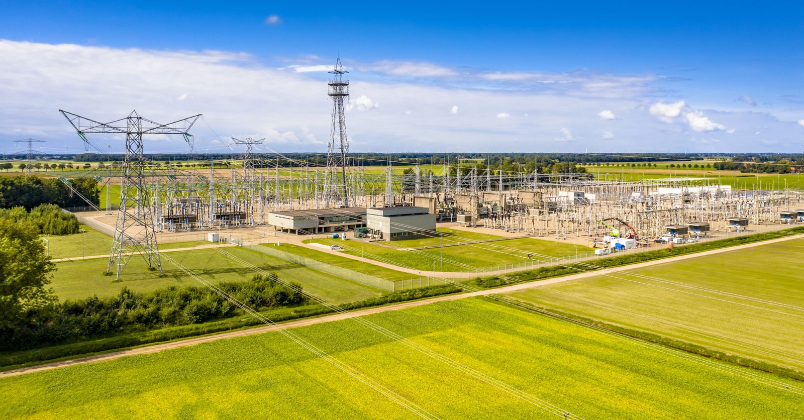 Aerial view of a large electrical substation with metal lattice towers, transformers, and many power lines across green fields under a blue sky.